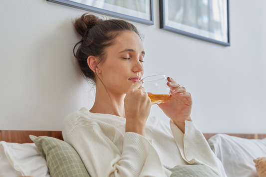 A woman sitting comfortably on a bed with cozy pillows, wearing a white sweater, and peacefully enjoying a cup of tea. She has her eyes closed, savoring the aroma of the warm tea in a clear glass cup, creating a serene and relaxing atmosphere.