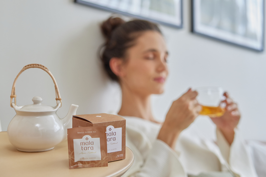 Woman peacefully drinking tea behind a ceramic teapot and Mala Tara Coffee Flower tea box and cup.