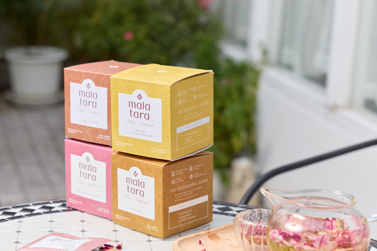 Outdoor display of Mala Tara organic flower tea boxes stacked, with teacup and flowers nearby.