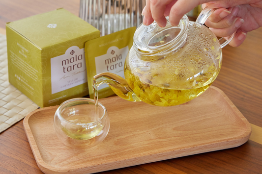 Hand pouring Mala Tara Yellow Chrysanthemum Tea into a small glass cup on wooden tray.