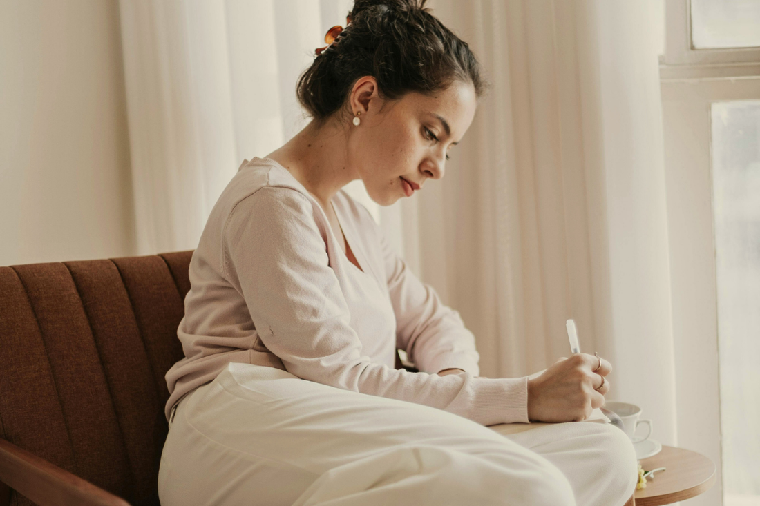 Woman writing in a notebook beside a cup of tea in a calm, sunlit room.